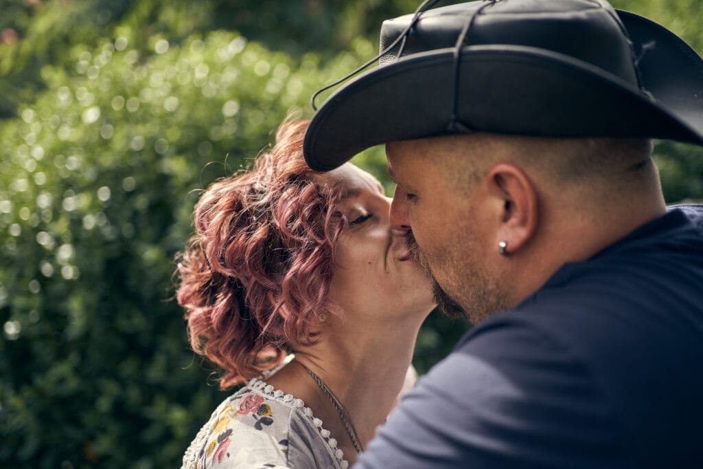 Couple échangeant un baiser tendre en extérieur, la femme avec des cheveux bouclés rose pastel et une robe à motifs floraux, et l'homme portant un chapeau de style cowboy, sur un arrière-plan flou de verdure.