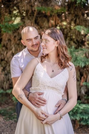 Couple attendant un enfant, l'homme tenant tendrement la femme enceinte par la taille, vêtue d'une robe blanche en dentelle de mariage, posant dans un jardin verdoyant et lumineux.